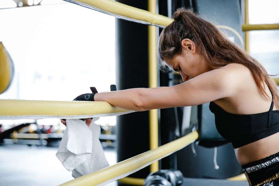 Kobieta w stroju sportowym opiera się o żółte liny ringu bokserskiego.