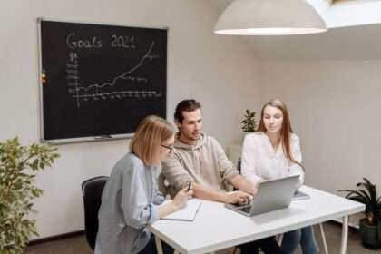 Trzy osoby pracują wspólnie przy laptopie w biurze z tablicą w tle.