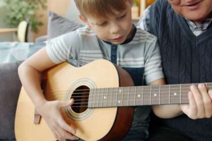 Starszy mężczyzna uczy chłopca grać na gitarze akustycznej siedząc na kanapie.