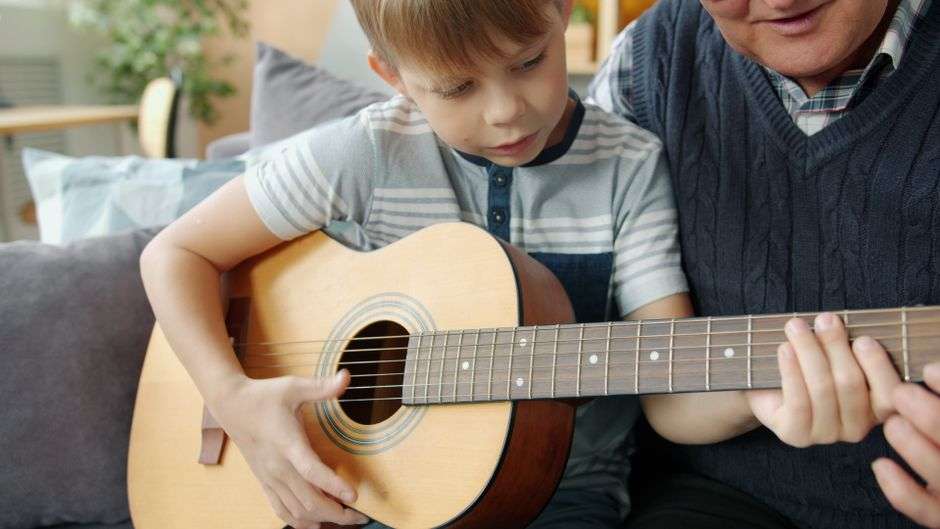 Starszy mężczyzna uczy chłopca grać na gitarze akustycznej siedząc na kanapie.