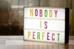 Podświetlana tablica z kolorowym napisem NOBODY IS PERFECT stojąca na stole.