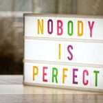 Podświetlana tablica z kolorowym napisem NOBODY IS PERFECT stojąca na stole.