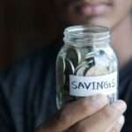 Mężczyzna trzyma szklany słoik wypełniony monetami z naklejką „SAVINGS”.