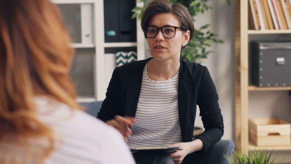 Kobieta w okularach i marynarce rozmawia z drugą osobą podczas spotkania.