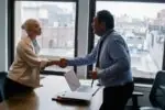 Kobieta i mężczyzna w biurze wykonują gest uścisku dłoni nad biurkiem.