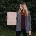 Kobieta w szarym swetrze trzyma karton z napisem "LOVE SHOULDN'T HURT".