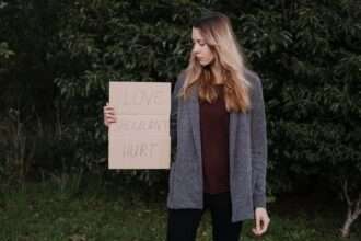 Kobieta w szarym swetrze trzyma karton z napisem "LOVE SHOULDN'T HURT".