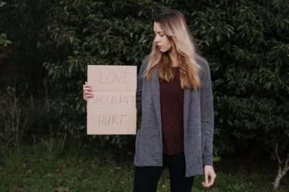 Kobieta w szarym swetrze trzyma karton z napisem "LOVE SHOULDN'T HURT".