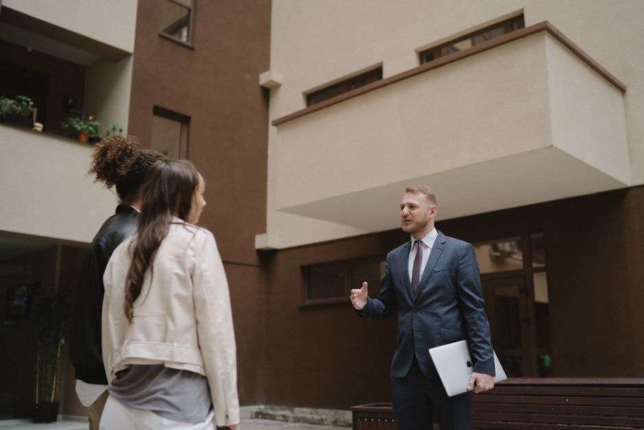 Mężczyzna w garniturze rozmawia z dwiema kobietami przed nowoczesnym budynkiem mieszkalnym.